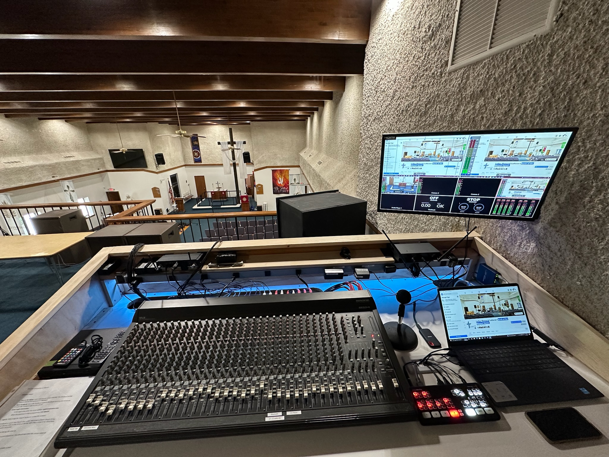 Close-up of a church’s AV control station: faders and knobs on the mixer alongside a livestream system, overlooking the worship space.